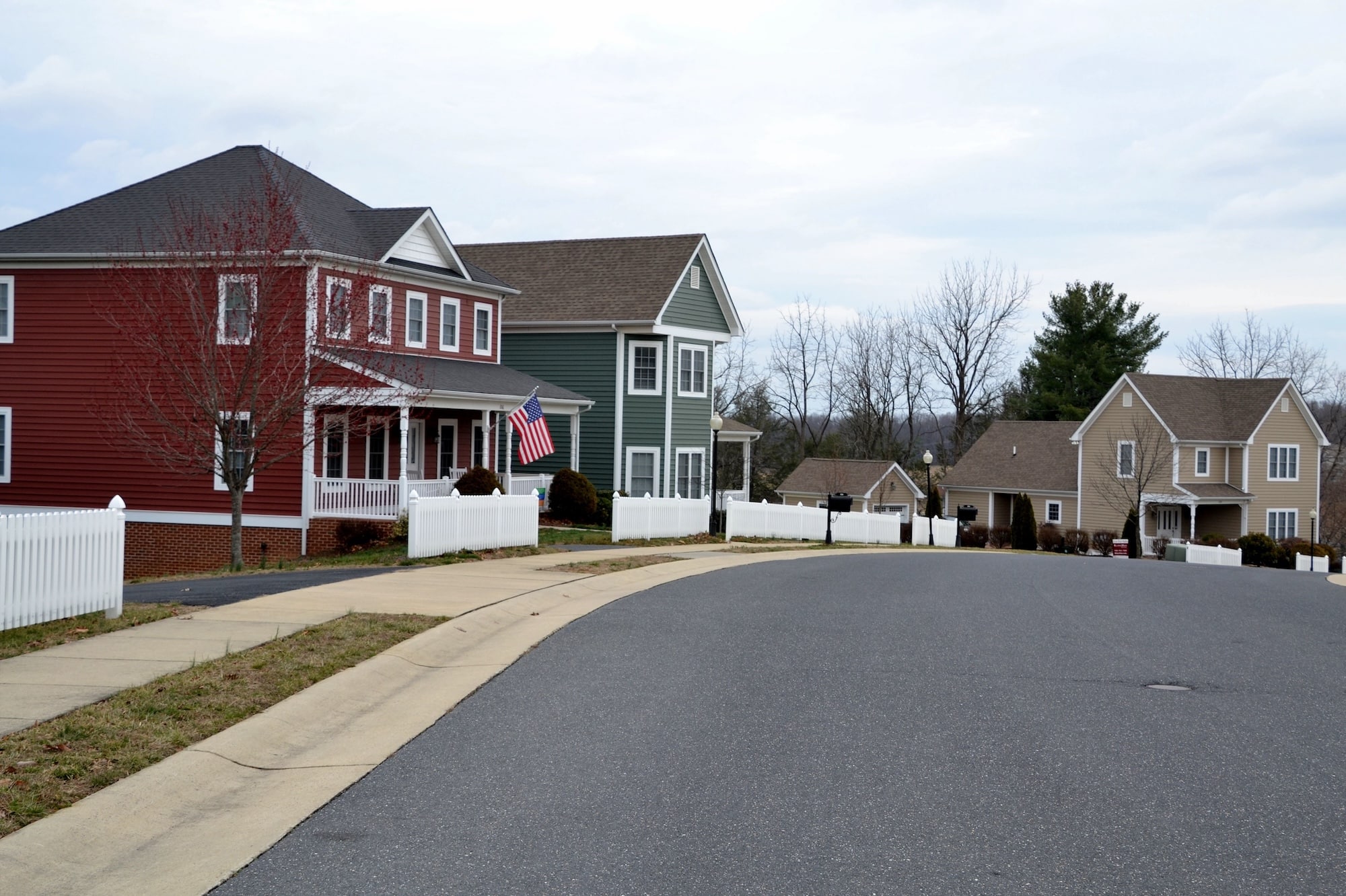 Suburban street with two-story houses in red, green, and beige. White picket fences line the yards. The sky is overcast, creating a calm atmosphere.