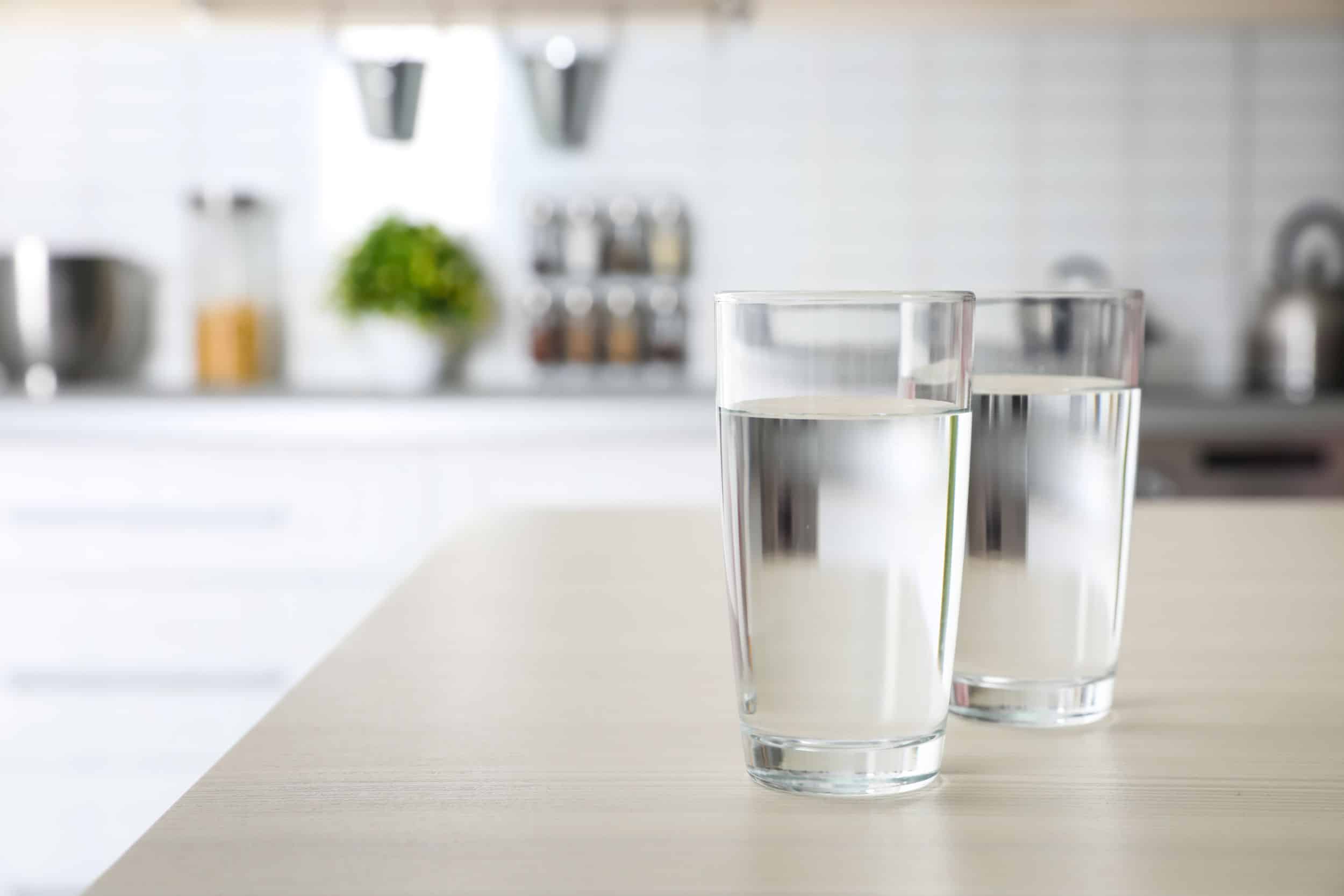 Two glasses of clear, fresh water sit on a light wooden table in a bright, modern kitchen.