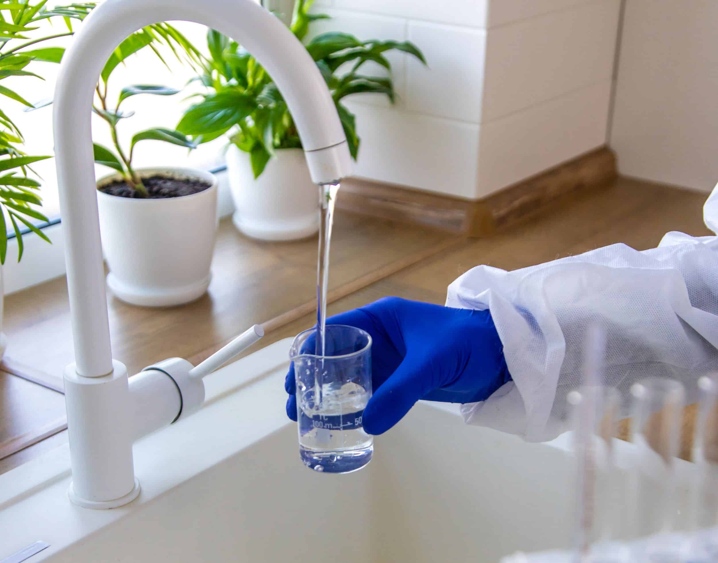 A technician wearing protective gear and blue gloves collects a water sample in a glass beaker from a white kitchen faucet for laboratory analysis.