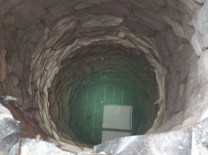 A photograph looking directly down into a deep, stone-lined circular well with clear water at the bottom reflecting light.