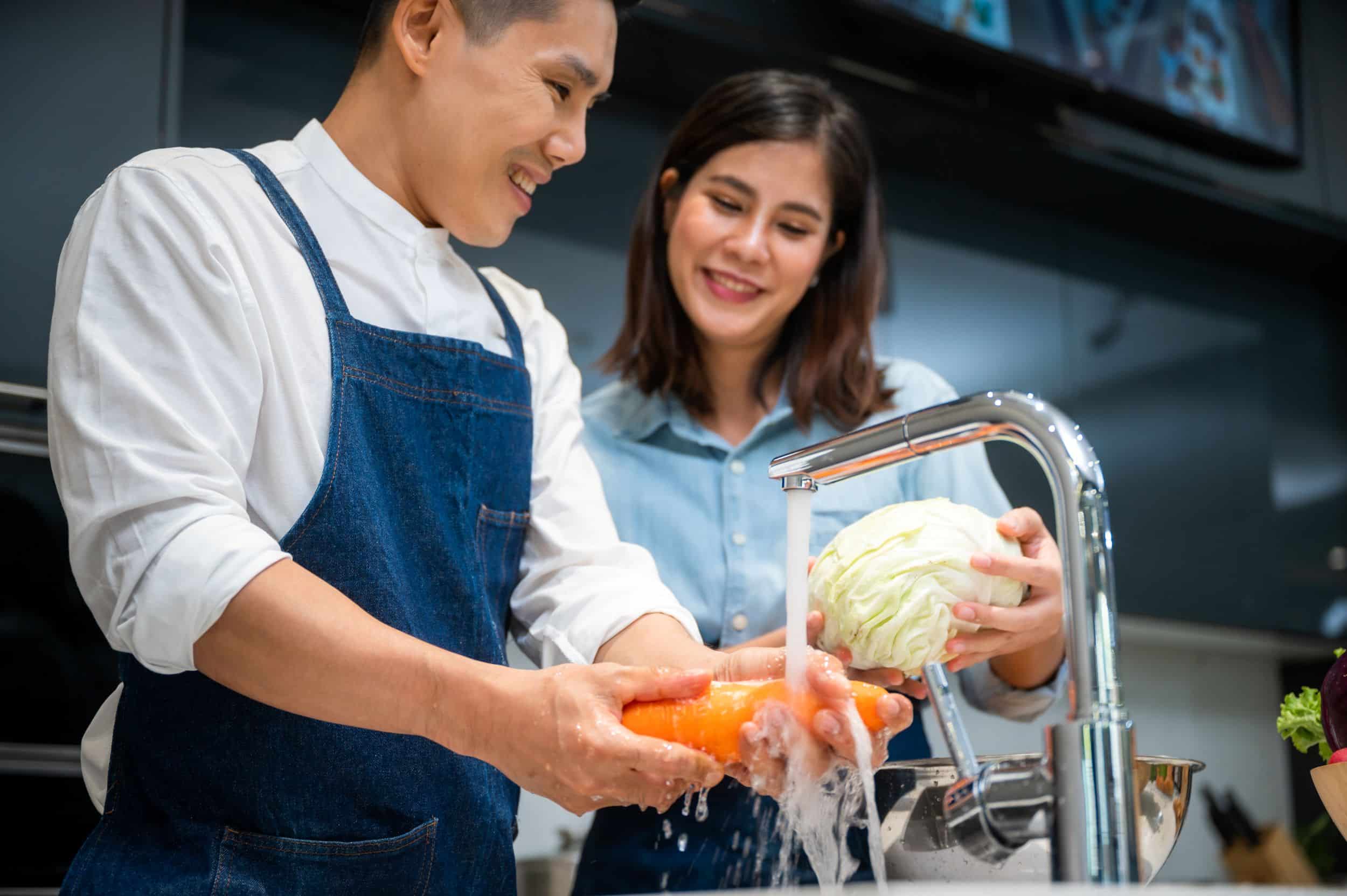 A smiling man and woman wash fresh vegetables, including a carrot and a head of cabbage, under a running kitchen faucet.