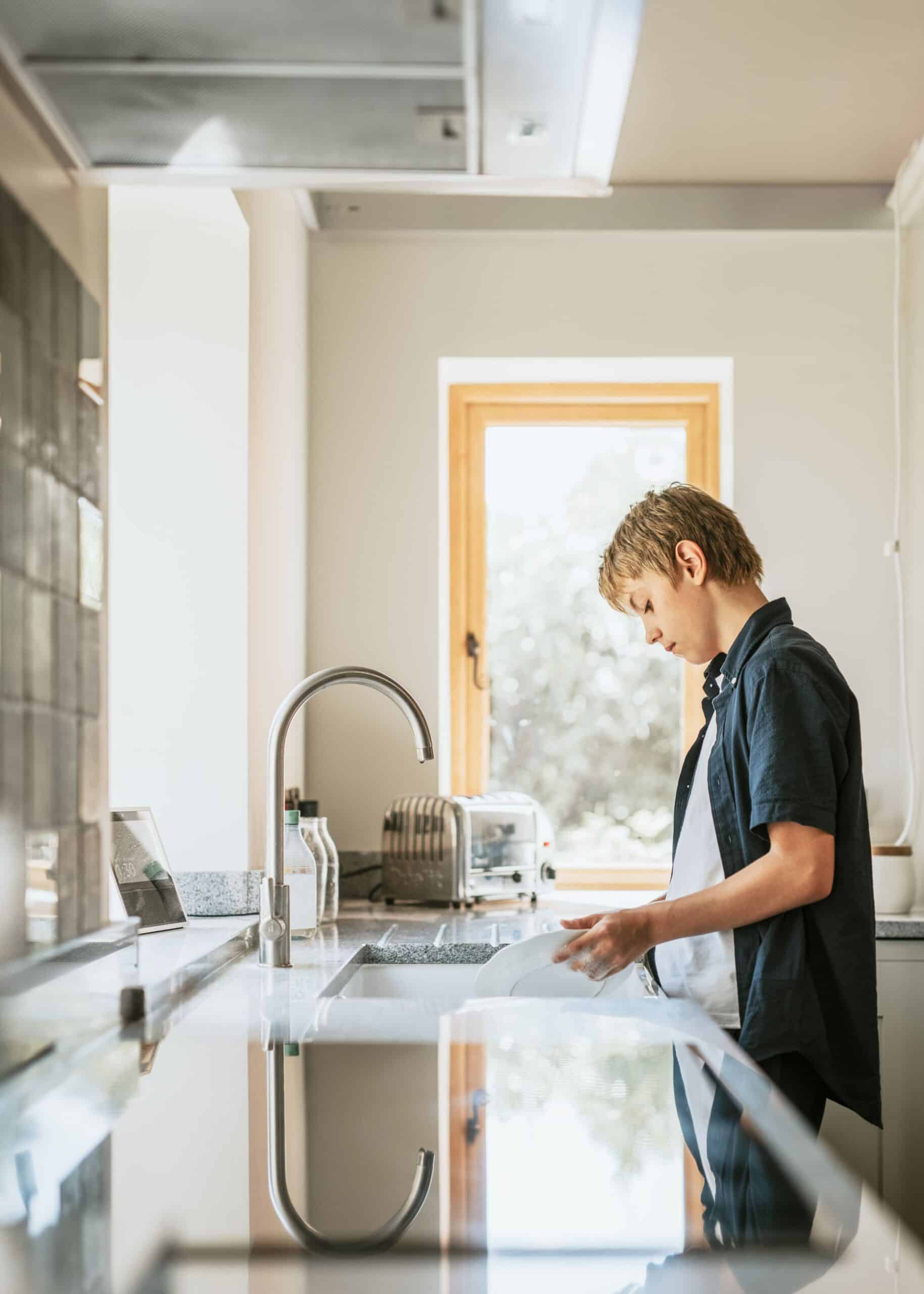 A young boy washes a white plate in a modern kitchen sink in front of a bright window.