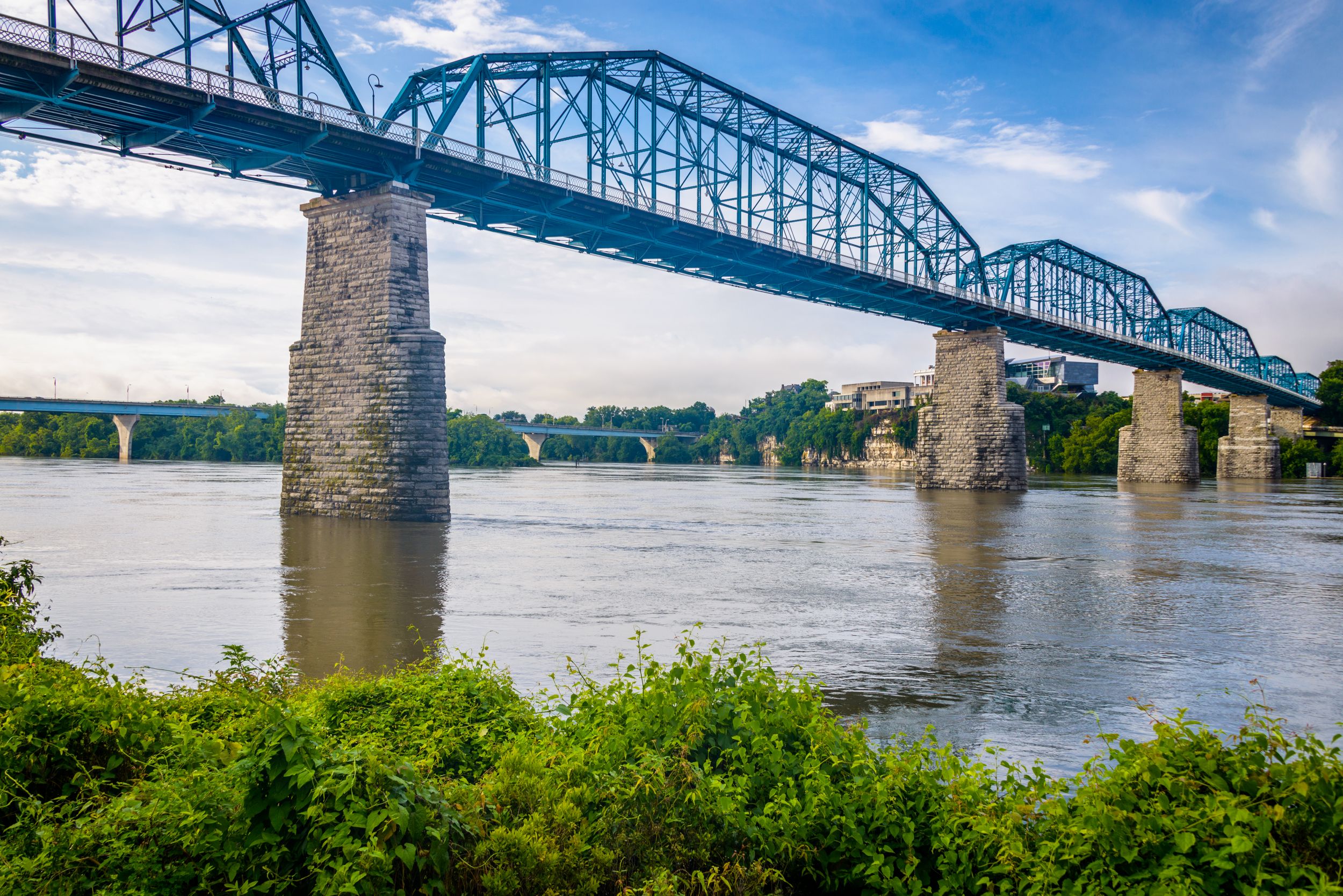A historic blue steel truss bridge with massive stone piers spans a wide river, seen from a riverbank with lush green foliage under a bright, cloudy sky.