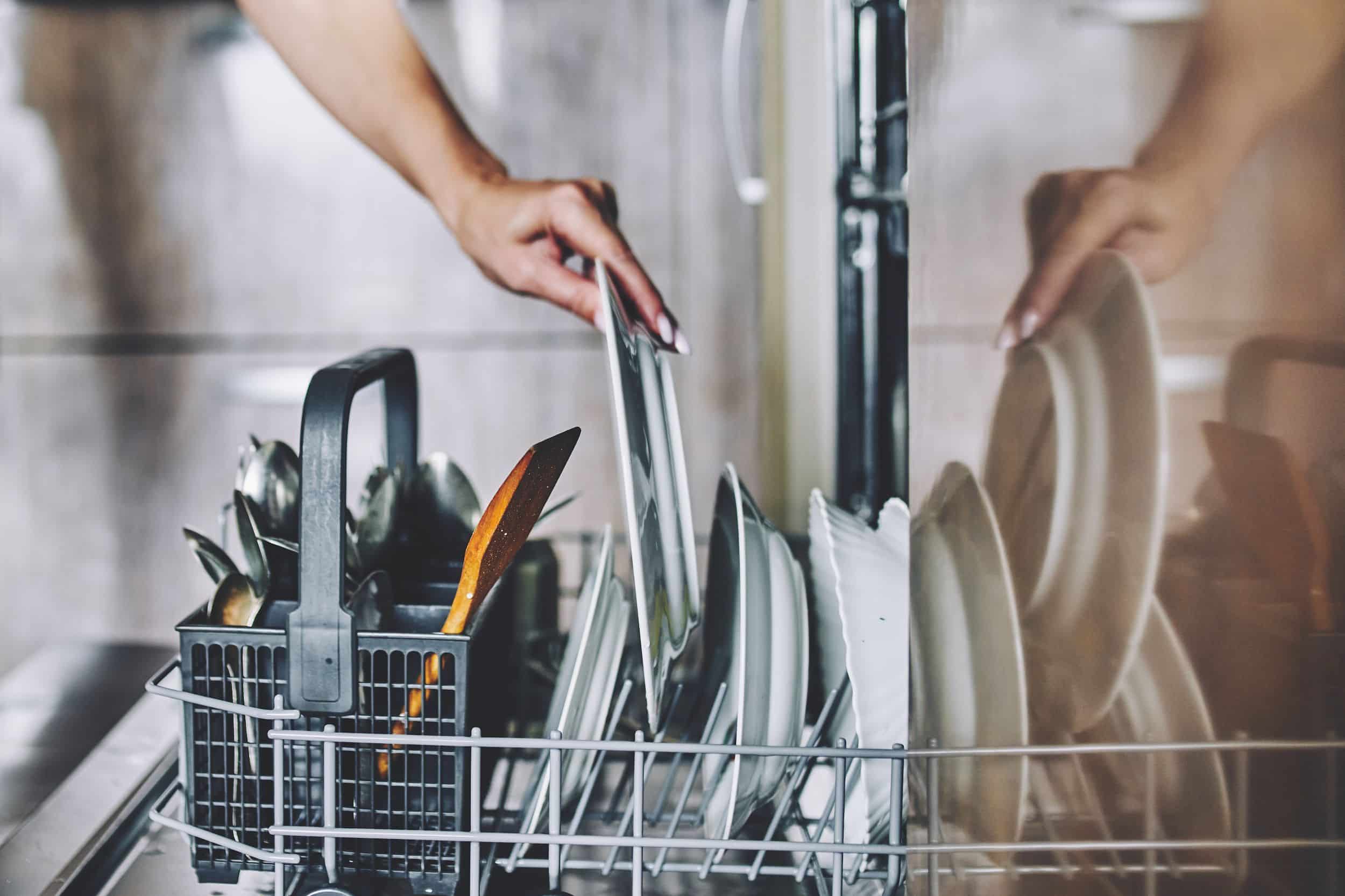 A person’s hand loads a white plate into a dishwasher rack filled with dishes and a cutlery basket.