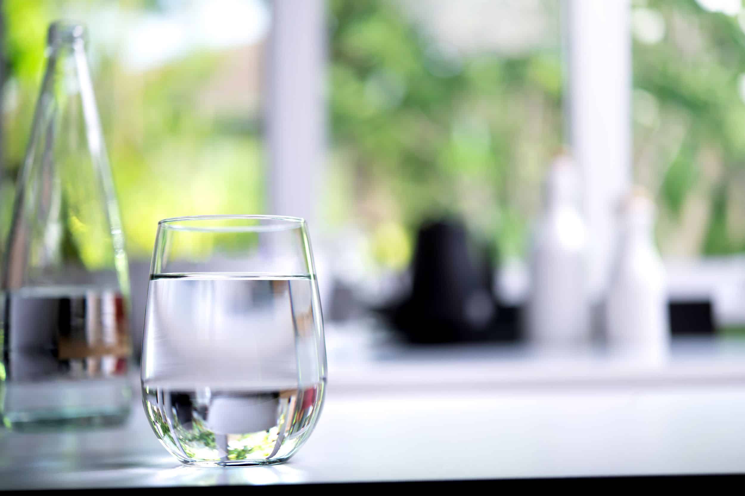 A clear glass and bottle of water sit on a white surface in a bright room with a blurred garden view in the background.