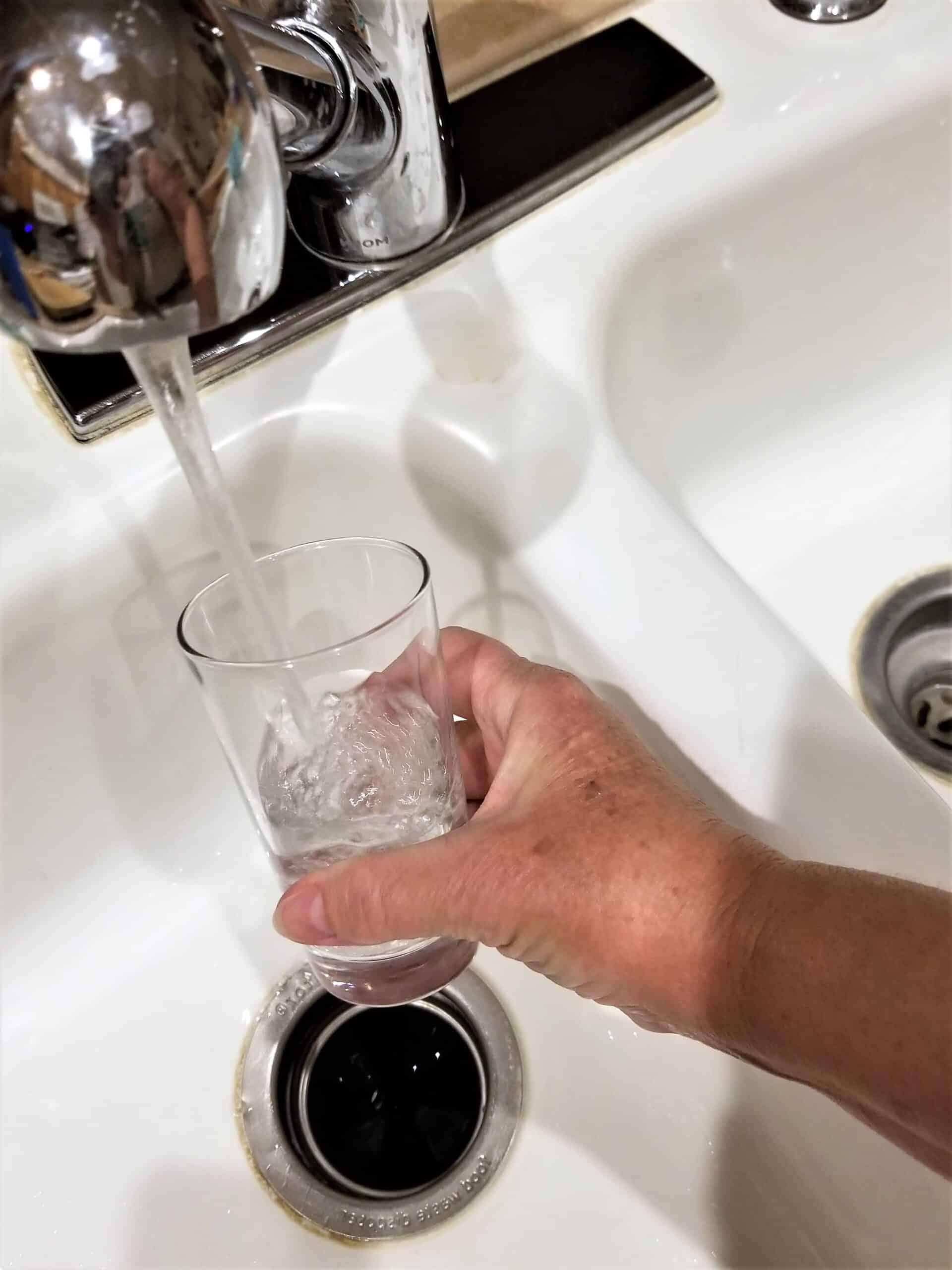 A hand holds a clear glass under a running chrome faucet, filling it with water over a white kitchen sink.