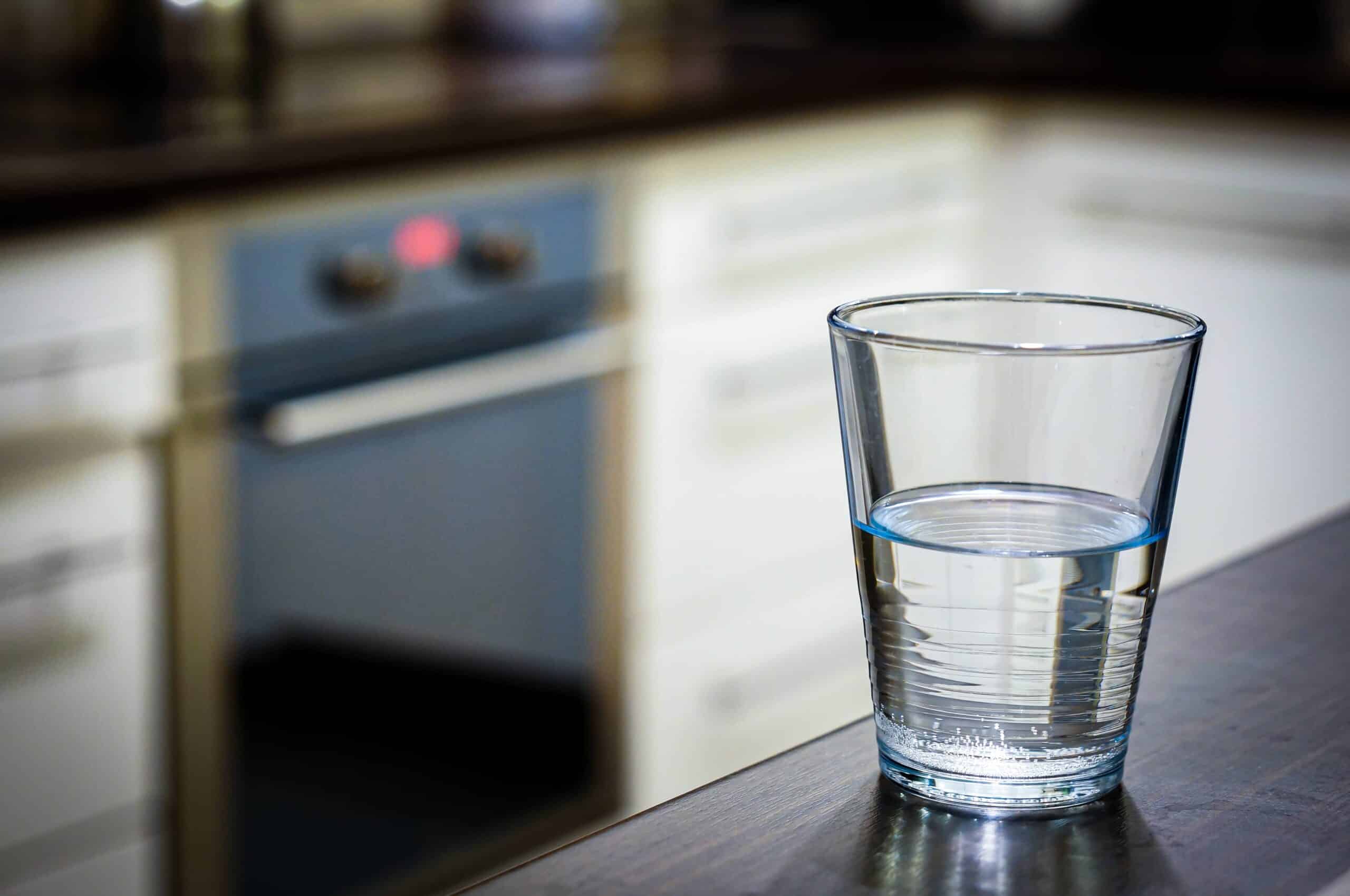 A clear glass half-filled with water sits on a dark countertop with a blurred kitchen background featuring an oven and white cabinets.