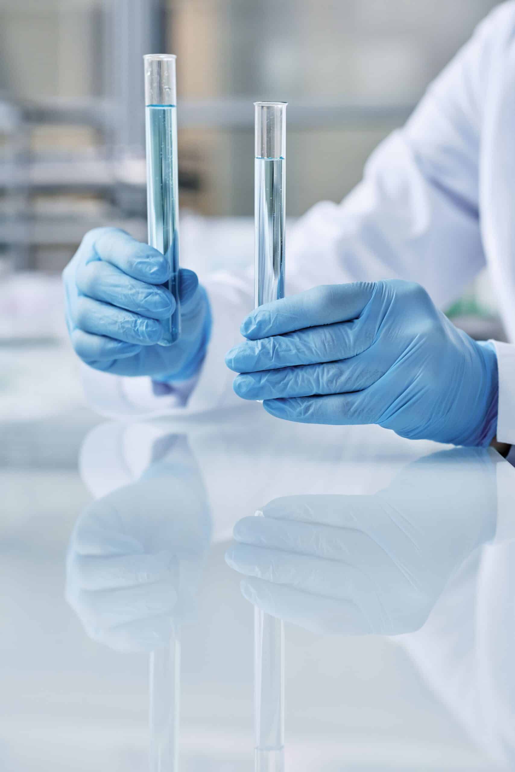 A person wearing a lab coat and blue gloves holds two test tubes filled with light blue liquid upright on a reflective white laboratory surface.