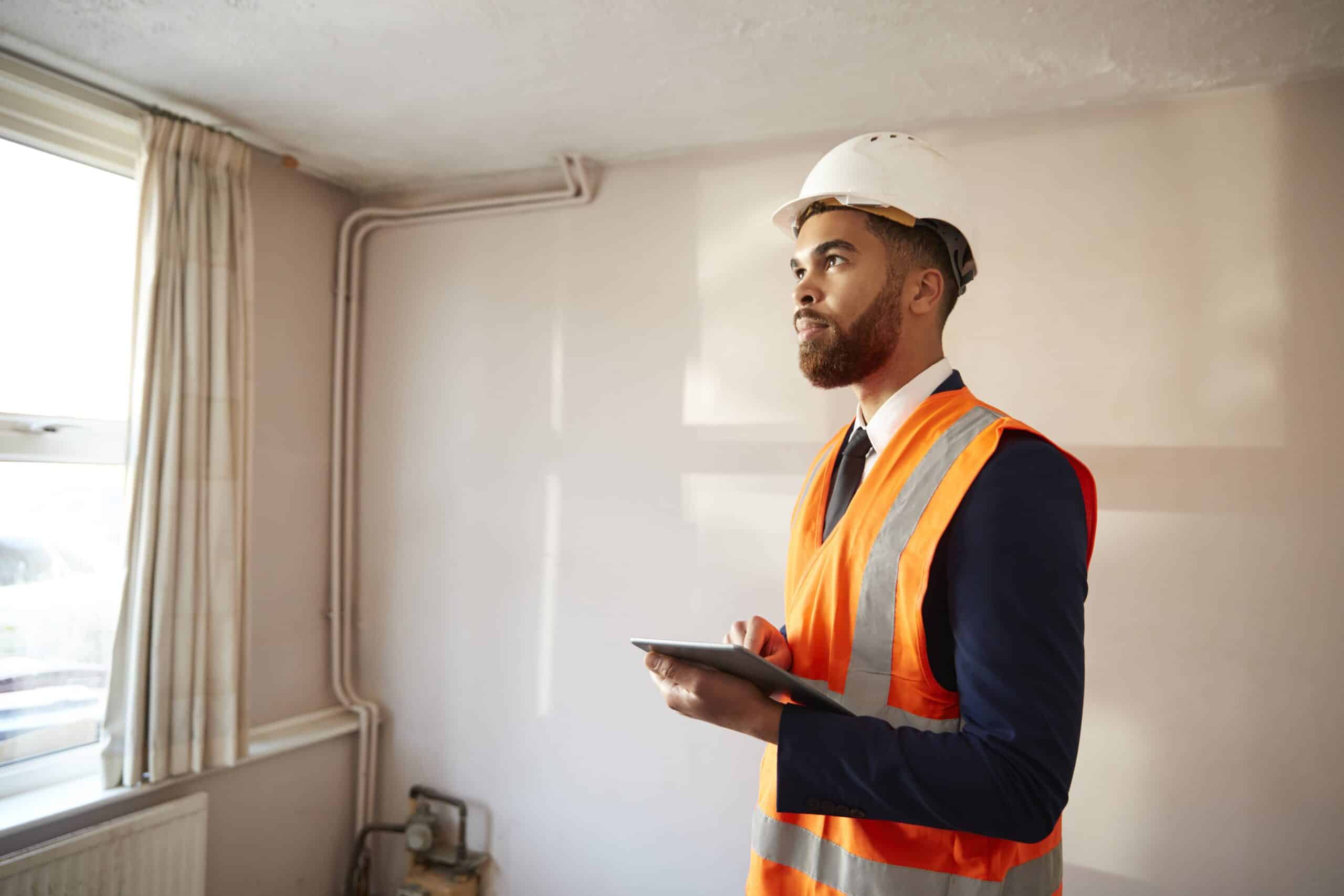 A man in a white hard hat and orange safety vest holds a tablet while inspecting an indoor room with exposed piping and natural light.