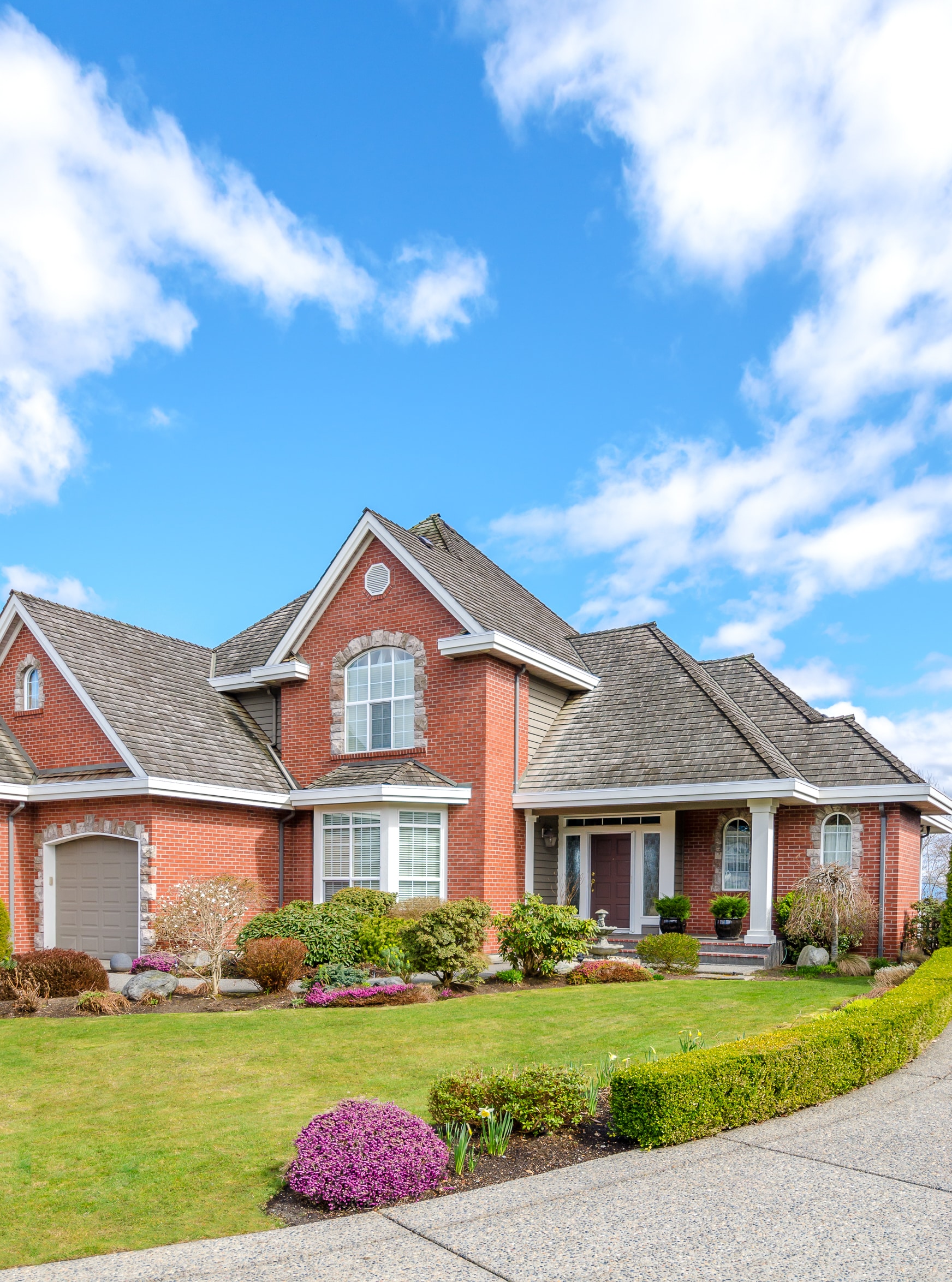 A large red brick house with a multi-gabled grey roof features a manicured green lawn and landscaped garden under a bright blue sky with scattered clouds.