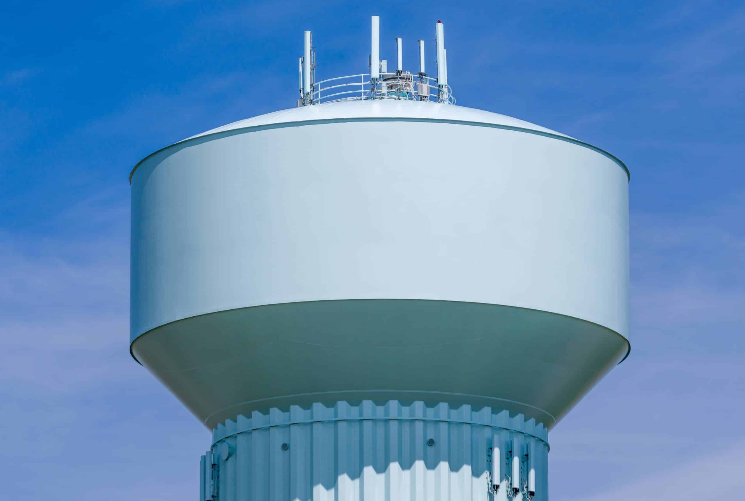 A large, light blue water tower with several telecommunications antennas mounted on top stands against a clear blue sky.