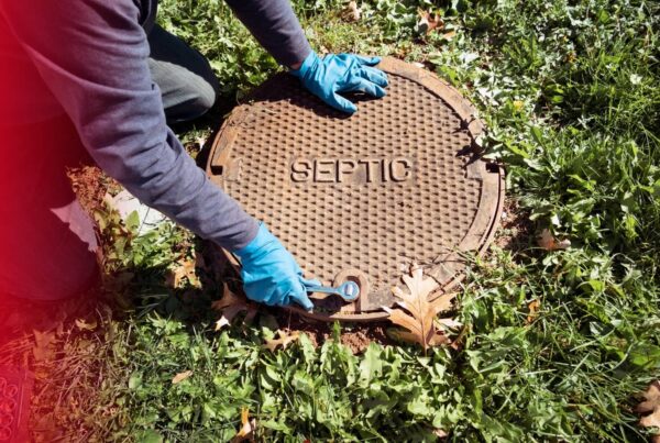 A person wearing blue gloves uses a wrench to open a heavy metal septic tank cover on a grassy lawn.