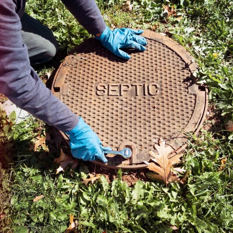 A person wearing blue gloves uses a wrench to open a heavy metal septic tank cover on a grassy lawn.