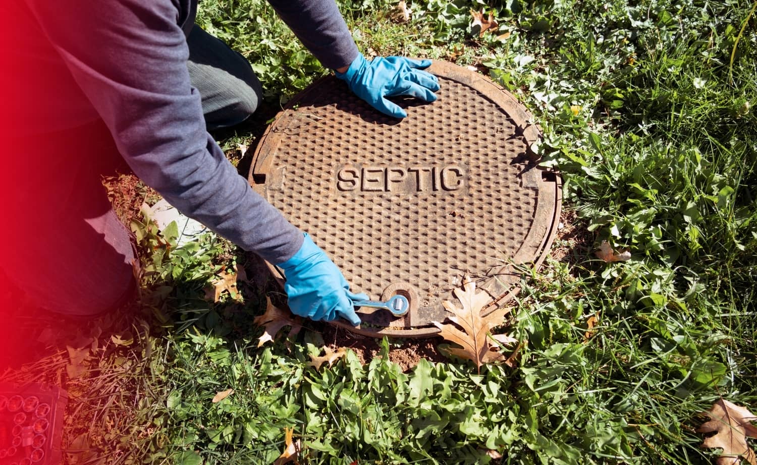 A person wearing blue gloves uses a wrench to open a heavy metal septic tank cover on a grassy lawn.