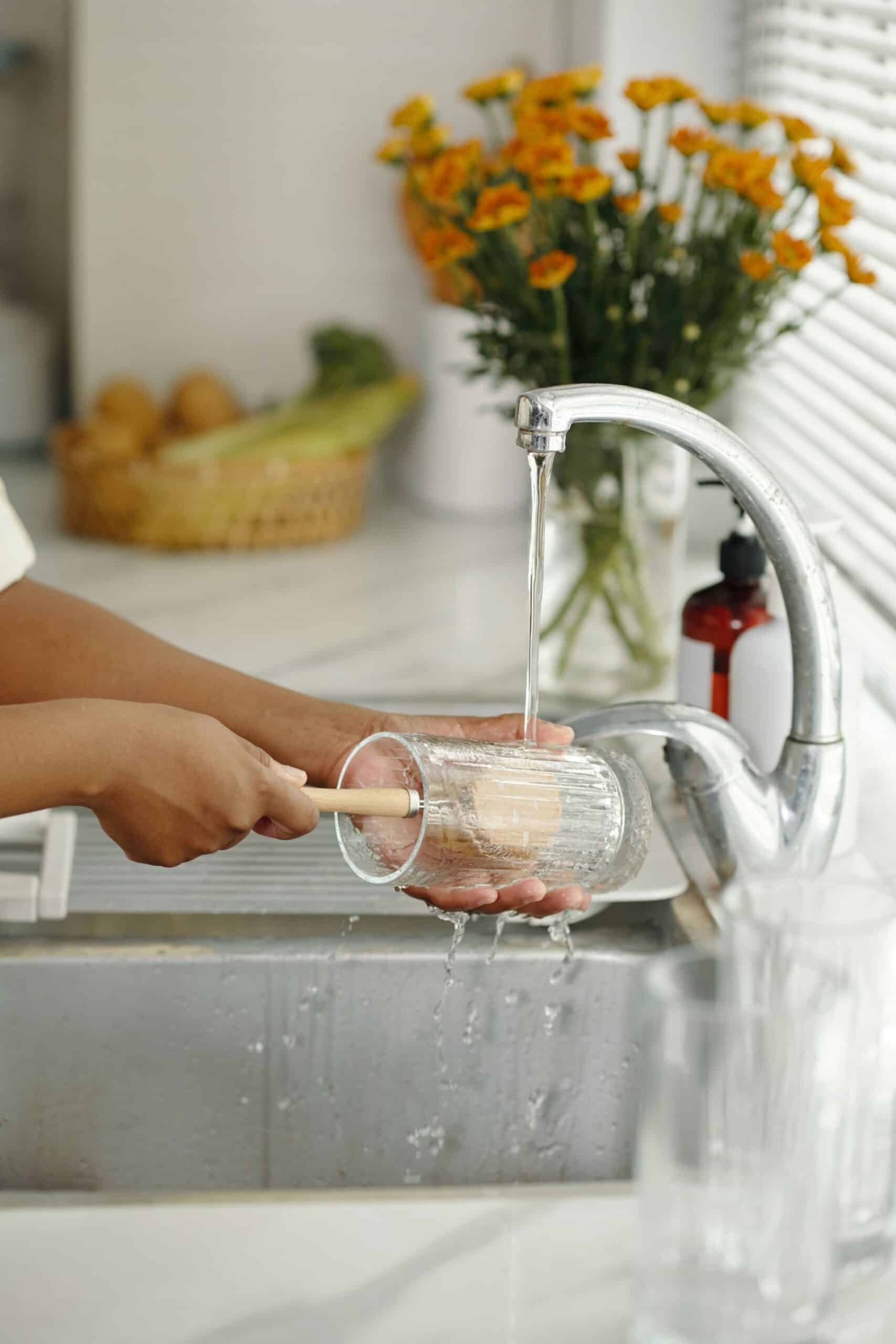 A person cleans a ribbed glass with a wooden brush under running water in a bright kitchen featuring orange flowers and a window with blinds.