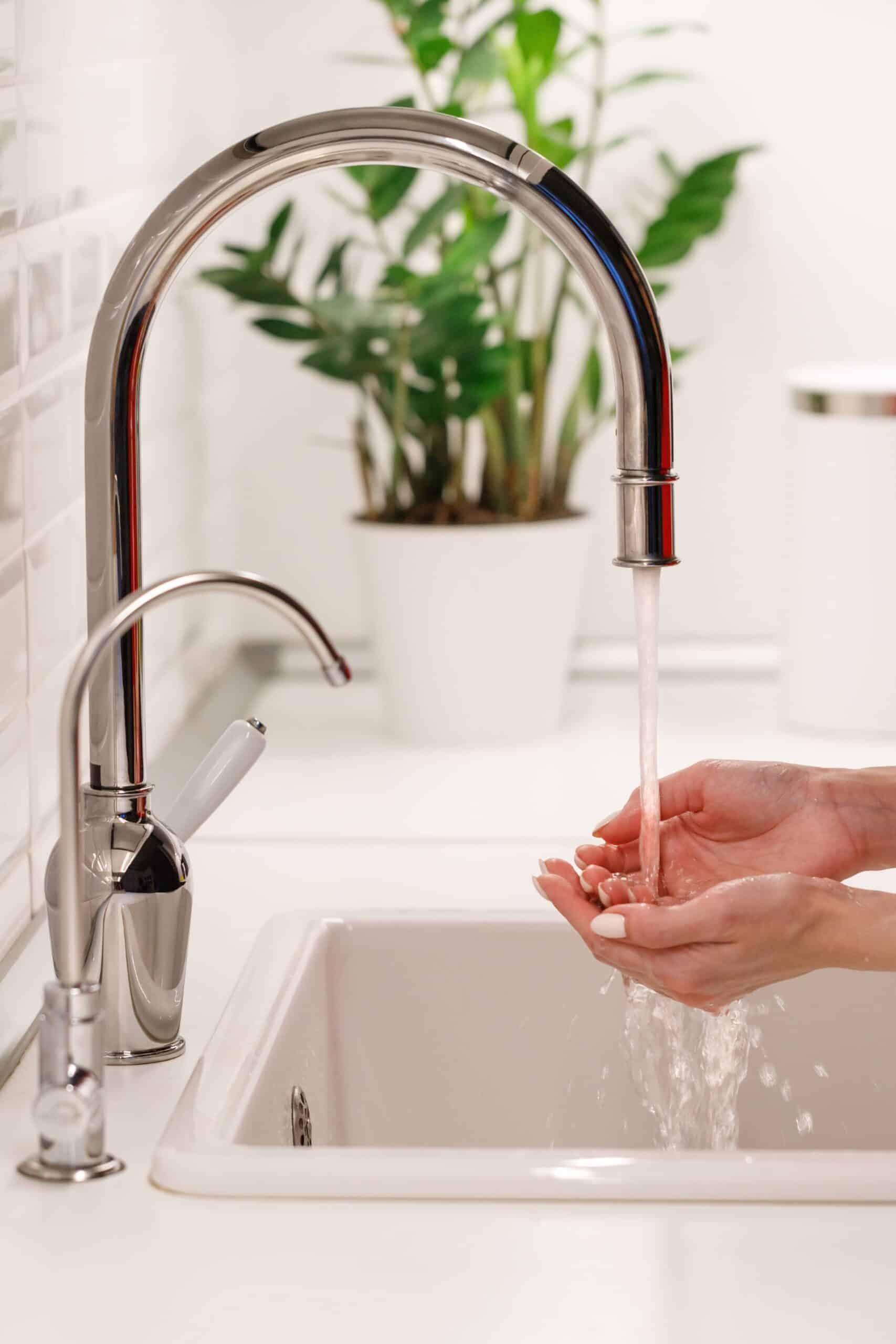 A person's hands are cupped under a stream of flowing water from a tall chrome faucet in a clean, white kitchen sink