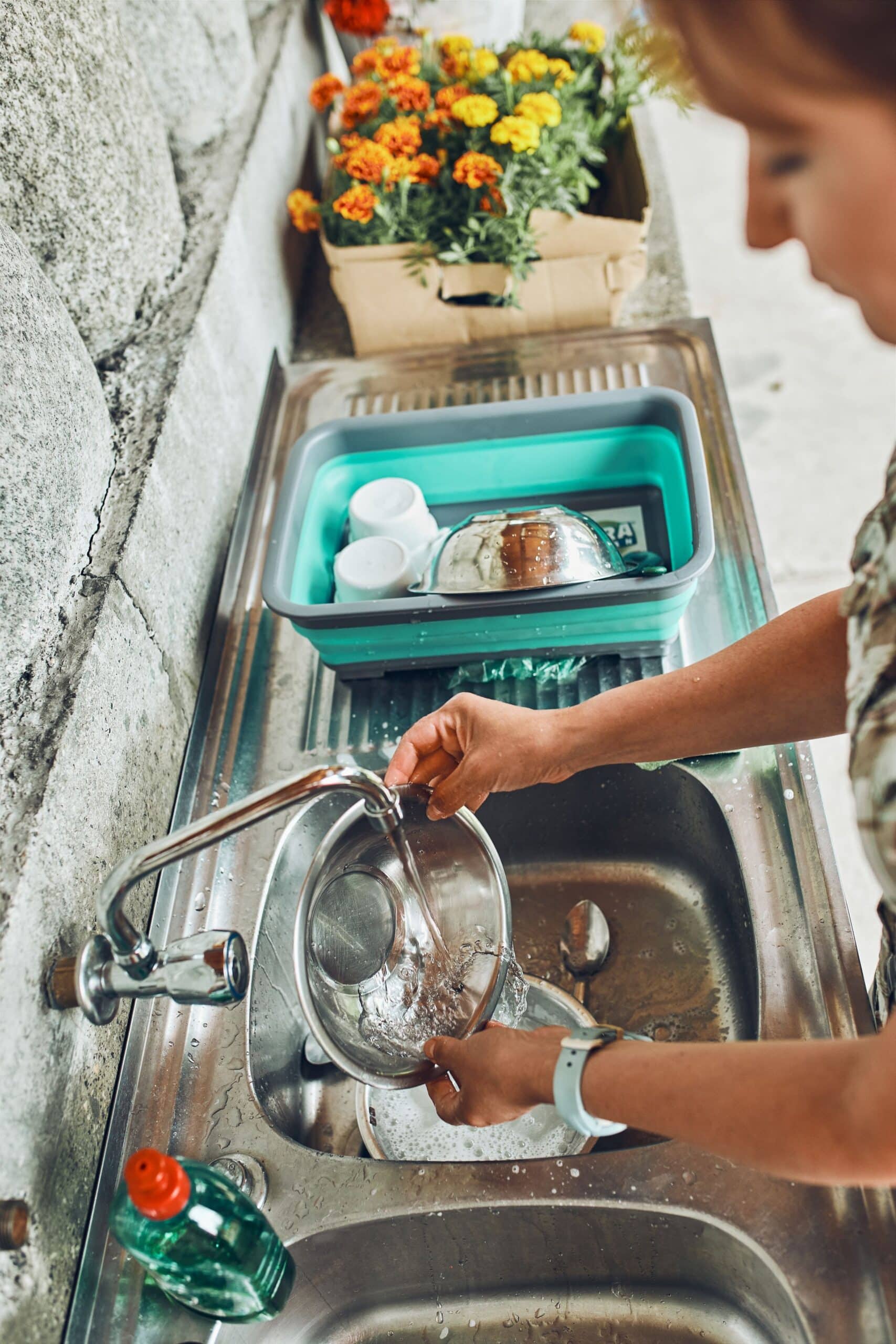 A woman rinses a metal bowl under a running faucet in a stainless steel sink, with a turquoise dish rack and orange flowers in the background.