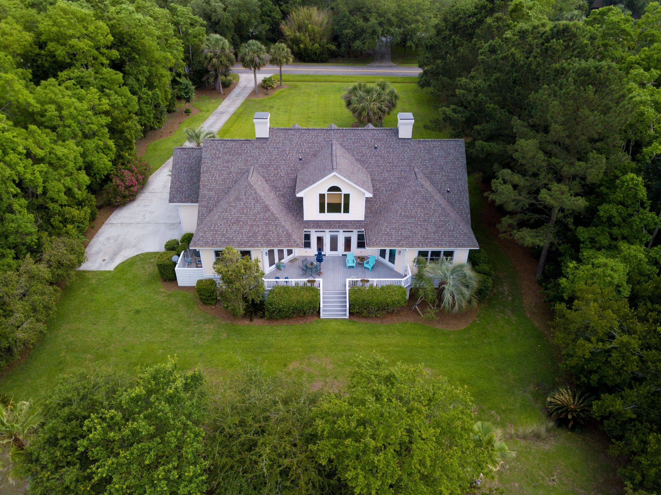 An aerial view of a large, two-story home with a newly shingled roof and a spacious back deck, nestled on a vibrant green lawn surrounded by dense woods.