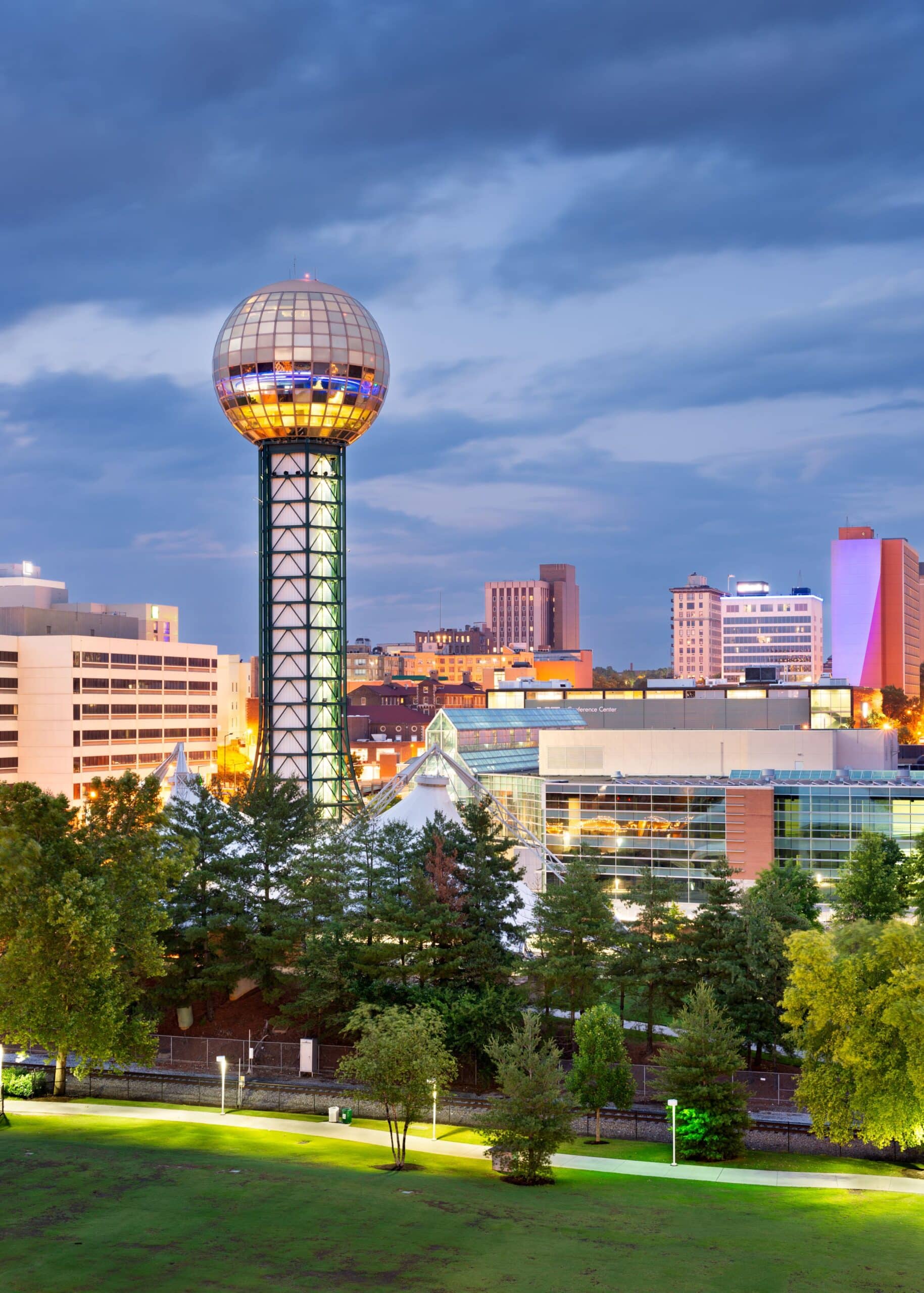 The iconic gold-sphered Sunsphere tower stands prominently against the Knoxville, Tennessee skyline at twilight, overlooking illuminated city buildings and a green park.